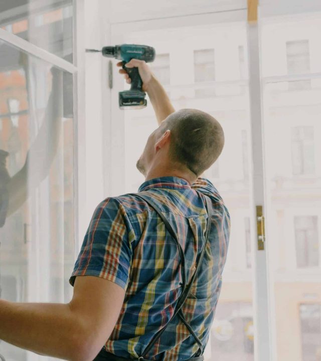 Back view of worker in checkered shirt drilling window frame during renovation process in apartment